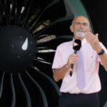 Man wearing a pink shirt and purple tie stood holding a microphone in front of aircraft engine